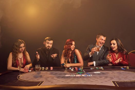 Merry partners are playing poker at casino. They are making bets waiting for a big win while posing sitting at the table against a dark smoke background in a ray of a spotlight. Cards, chips, money, alcohol, gambling, entertainment concept.