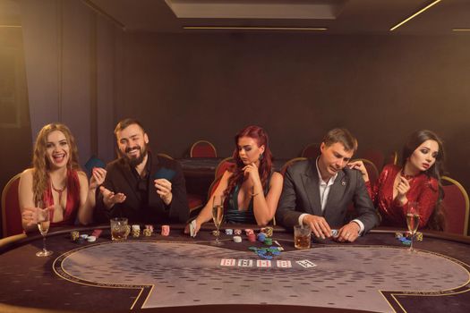 Merry colleagues are playing poker at casino. They are making bets waiting for a big win while posing at the table against a dark smoke background in a ray of a spotlight. Cards, chips, money, alcohol, gambling, entertainment concept.