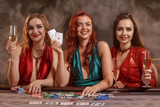 Gorgeous women are playing poker at casino. They are celebrating their win, smiling, looking at the camera and posing at the table against a a dark smoke background. Cards, chips, money, alcohol, gambling, entertainment concept.