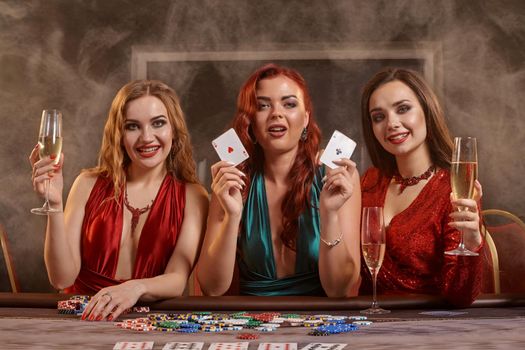 Charming girls are playing poker at casino. They are celebrating their win, smiling, looking at the camera and posing at the table against a a dark smoke background. Cards, chips, money, alcohol, gambling, entertainment concept.