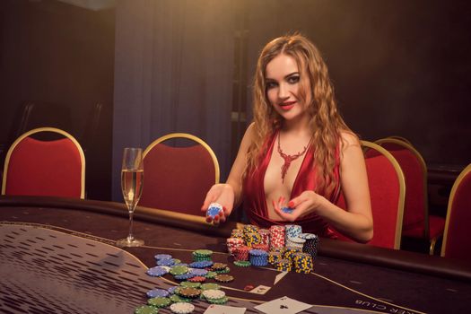 Pretty woman with a long curly hair and perfect make-up, dressed in a sexy red dress. She is rejoicing her win while sitting at a gambling table and looking at the camera. Poker concept on a dark smoke background in a ray of a yellow spotlight. Casino.