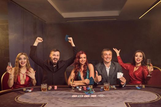 Optimistic colleagues are playing poker at casino. They are celebrating their win, smiling and looking vey excited while posing sitting at the table against a dark smoke background in a ray of a spotlight. Cards, chips, money, alcohol, gambling, entertainment concept.