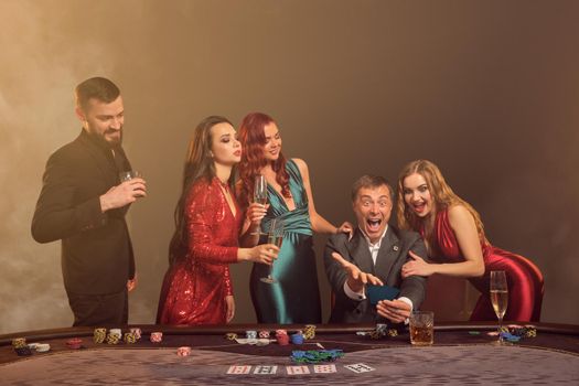 Funny friends are playing poker at casino. They are celebrating their win, smiling and looking vey excited while posing at the table against a dark smoke background in a ray of a spotlight. Cards, chips, money, alcohol, gambling, entertainment concept.