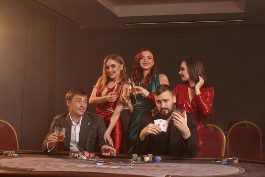 Cheerful buddies are playing poker at casino. They are celebrating their win, smiling and posing at the table against a dark smoke background. Cards, chips, money, alcohol, fortune, gambling, entertainment concept.