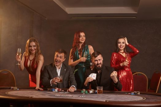 Cheerful friends are playing poker at casino. They are celebrating their win, smiling and posing at the table against a dark smoke background. Cards, chips, money, alcohol, fortune, gambling, entertainment concept.
