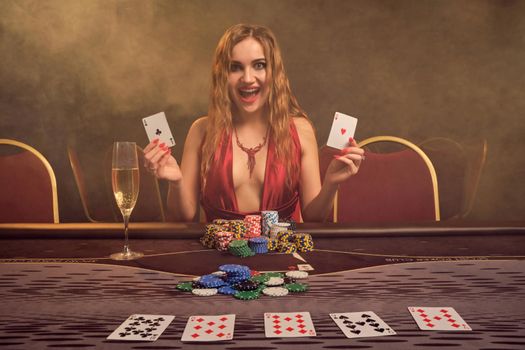 Alluring girl with a long curly hair and perfect make-up, dressed in a sexy red dress. She is sitting at a gambling table with two playing cards in her hands and looking at the camera. Poker concept on a dark smoke background in a ray of a yellow spotlight. Casino.