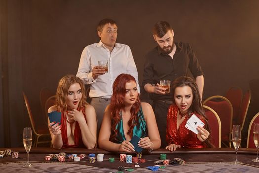 Agitated colleagues are playing poker at casino. They are celebrating their win, smiling and posing at the table against a dark smoke background. Cards, chips, money, alcohol, fortune, gambling, entertainment concept.