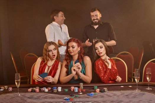 Agitated companions are playing poker at casino. They are celebrating their win, smiling and posing at the table against a dark smoke background. Cards, chips, money, alcohol, fortune, gambling, entertainment concept.