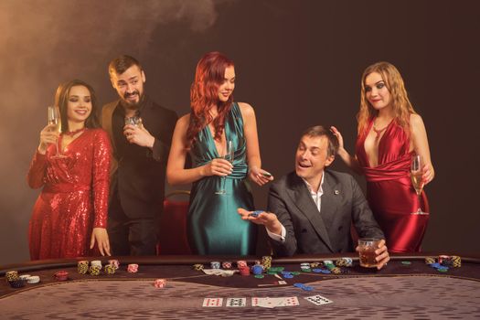 Happy classmates are playing poker at casino. They are celebrating their win, smiling and looking vey excited while posing at the table against a dark smoke background in a ray of a spotlight. Cards, chips, money, alcohol, gambling, entertainment concept.