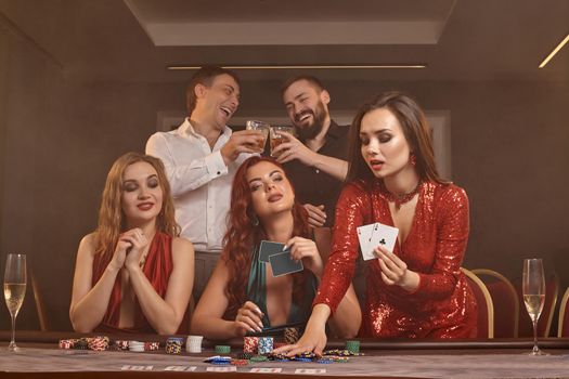 Group of an enthusiastic colleagues are playing poker at casino. They are celebrating their win, looking at the camera and holding some playing cards in their hands while posing at the table against a smoke background. Chips, money, alcohol, gambling, entertainment concept.