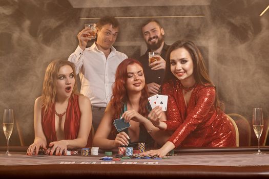 Happy companions are playing poker at casino. They are celebrating their win, smiling and posing at the table against a dark smoke background. Cards, chips, money, alcohol, fortune, gambling, entertainment concept.