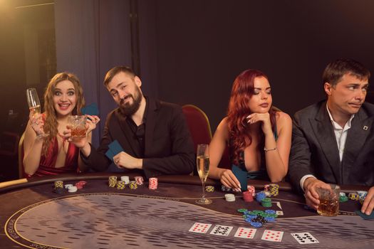 Merry friends are playing poker at casino. They are making bets waiting for a big win while posing sitting at the table against a dark smoke background in a ray of a spotlight. Cards, chips, money, alcohol, gambling, entertainment concept.