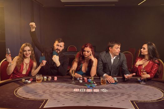 Merry classmates are playing poker at casino. They are making bets waiting for a big win while posing sitting at the table against a dark smoke background in a ray of a spotlight. Cards, chips, money, alcohol, gambling, entertainment concept.
