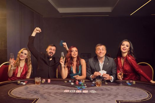 Optimistic buddies are playing poker at casino. They are celebrating their win, smiling and looking vey excited while posing sitting at the table against a dark smoke background in a ray of a spotlight. Cards, chips, money, alcohol, gambling, entertainment concept.