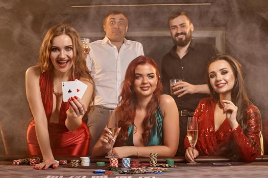 Happy classmates are playing poker at casino. They are celebrating their win, smiling and posing at the table against a dark smoke background. Cards, chips, money, alcohol, fortune, gambling, entertainment concept.
