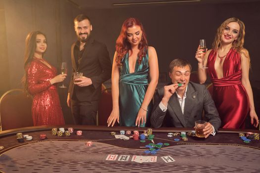 Jojful friends are playing poker at casino. They are celebrating their win, smiling and looking vey excited while posing at the table against a dark smoke background in a ray of a spotlight. Cards, chips, money, alcohol, gambling, entertainment concept.