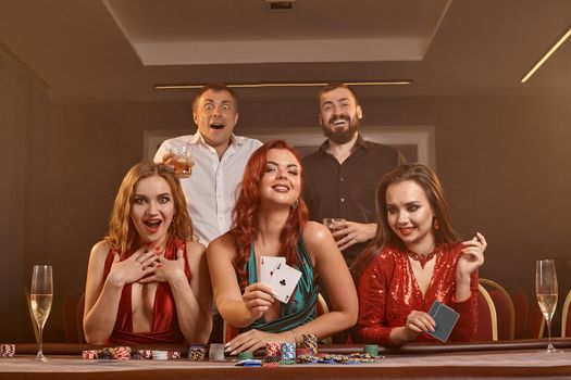 Group of an enthusiastic friends are playing poker at casino. They are celebrating their win, looking at the camera and holding some playing cards in their hands while posing at the table against a smoke background. Chips, money, alcohol, gambling, entertainment concept.