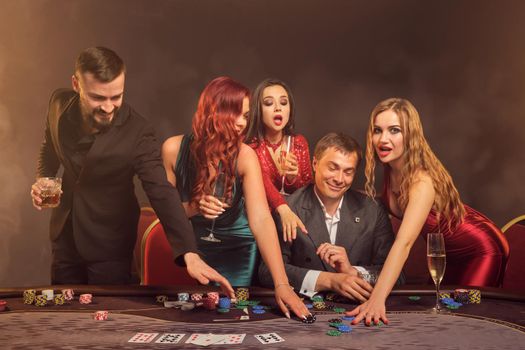 Happy friends are playing poker at casino. They are celebrating their win, smiling and looking vey excited while posing at the table against a dark smoke background in a ray of a spotlight. Cards, chips, money, alcohol, gambling, entertainment concept.