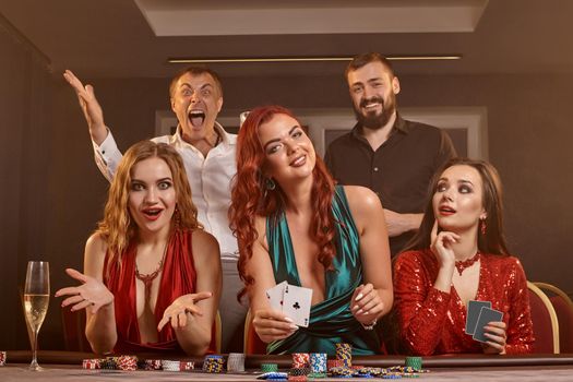 Group of a wealthy classmates are playing poker at casino. They are celebrating their win, looking at the camera and holding some playing cards in their hands while posing at the table against a smoke background. Chips, money, alcohol, gambling, entertainment concept.