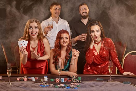 Fortunate friends are playing poker at casino. They are celebrating their win, smiling and posing at the table against a dark smoke background. Cards, chips, money, alcohol, gambling, entertainment concept.