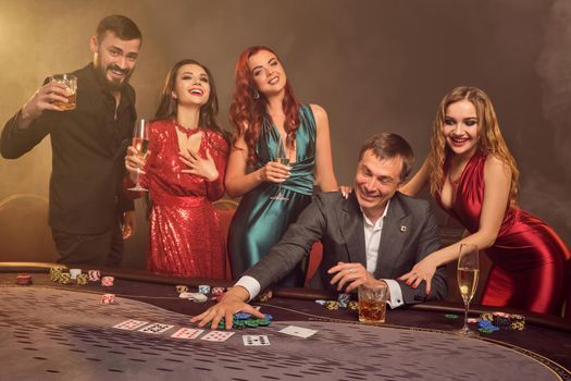 Enthusiastic colleagues are playing poker at casino. They are celebrating their win, smiling and looking vey excited while posing at the table against a dark smoke background in a ray of a spotlight. Cards, chips, money, alcohol, gambling, entertainment concept.