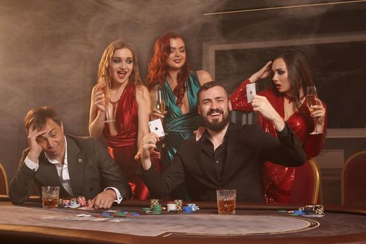 Excited colleagues are playing poker at casino. They are celebrating their win, smiling and posing at the table against a dark smoke background. Cards, chips, money, alcohol, fortune, gambling, entertainment concept.