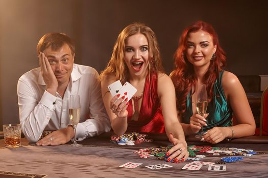 Excited classmates are playing poker at casino. They are celebrating their win, smiling and posing at the table against a dark smoke background. Cards, chips, money, alcohol, fortune, gambling, entertainment concept.