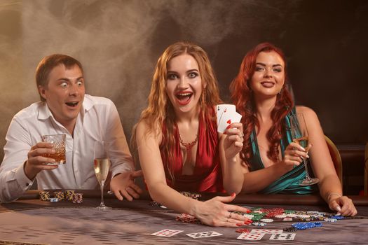 Group of a young wealthy friends are playing poker at casino. They are celebrating their win, looking at the camera and smiling while showing the winning combination and posing sitting at the table against a dark smoke background. Cards, chips, money, gambling, entertainment concept.