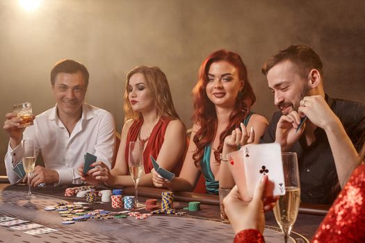 Pleasant friends are playing poker at casino. Youth are making bets waiting for a big win while posing at the table against a white spotlight on a dark smoke background. Cards, chips, money, alcohol, gambling, entertainment concept.