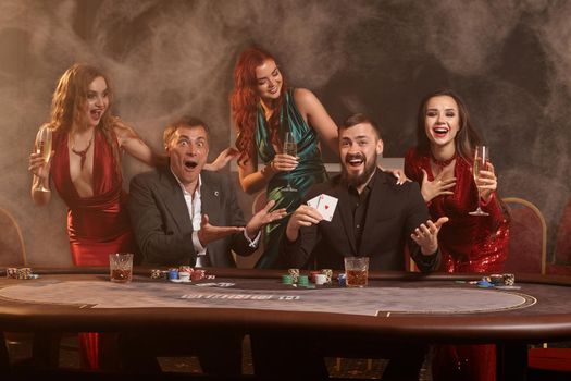 Cheerful companions are playing poker at casino. They are celebrating their win, smiling and posing at the table against a dark smoke background. Cards, chips, money, alcohol, fortune, gambling, entertainment concept.