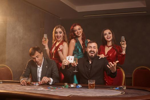 Cheerful classmates are playing poker at casino. They are celebrating their win, smiling and posing at the table against a dark smoke background. Cards, chips, money, alcohol, fortune, gambling, entertainment concept.