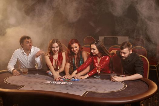 Wealthy friends are playing poker at casino. They are celebrating their win, smiling and looking vey excited while posing at the table against a dark smoke background. Cards, chips, money, alcohol, gambling, entertainment concept.