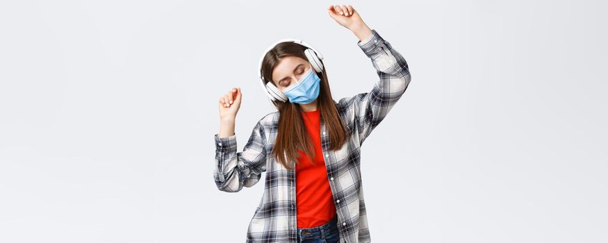 Social distancing, leisure and lifestyle on covid-19 outbreak, coronavirus concept. Carefree, relaxed good-looking woman in medical mask listening music in headphones, dancing with closed eyes.