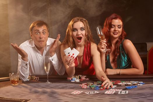Group of a young colleagues are playing poker at casino. They are celebrating their win, looking at the camera and smiling while showing the winning combination and posing sitting at the table against a dark smoke background. Cards, chips, money, gambling, entertainment concept.