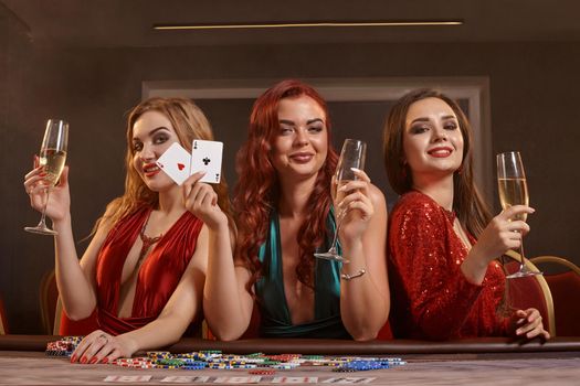 Three charming maidens are playing poker at casino. They are celebrating their win, looking at the camera and smiling, holding champagne and cards in their hands while posing at the table against a smoke background. Chips, money, gambling, entertainment concept.