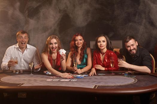 Cheerful buddies are playing poker at casino. They are celebrating their win, smiling and looking vey excited while posing at the table against a dark smoke background. Cards, chips, money, alcohol, gambling, entertainment concept.