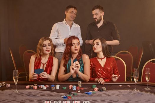Concentrated classmates are playing poker at casino. Golden youth are making bets waiting for a big win while posing at the table against a dark smoke background. Cards, chips, money, fortune, alcohol, gambling, entertainment concept.