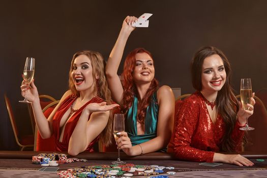 Three charming females are playing poker at casino. They are celebrating their win, looking at the camera and smiling, holding champagne and cards in their hands while posing at the table against a smoke background. Chips, money, gambling, entertainment concept.