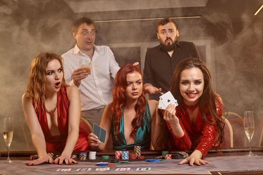 Joyful companions are playing poker at casino. They are celebrating their win, smiling and posing at the table against a dark smoke background. Cards, chips, money, alcohol, fortune, gambling, entertainment concept.