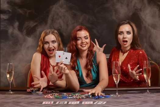 Attractive girls are playing poker at casino. They are celebrating their win, smiling, looking at the camera and posing at the table against a a dark smoke background. Cards, chips, money, alcohol, gambling, entertainment concept.