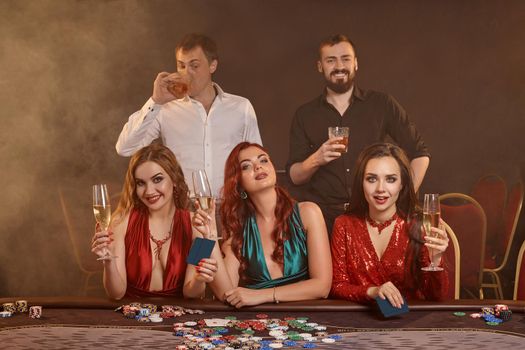 Enthusiastic classmates are playing poker at casino. They are celebrating their win, smiling and posing at the table against a dark smoke background. Cards, chips, money, alcohol, fortune, gambling, entertainment concept.