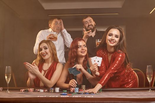 Happy colleagues are playing poker at casino. They are celebrating their win, smiling and posing at the table against a dark smoke background. Cards, chips, money, alcohol, fortune, gambling, entertainment concept.