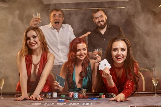 Joyful colleagues are playing poker at casino. They are celebrating their win, smiling and posing at the table against a dark smoke background. Cards, chips, money, alcohol, fortune, gambling, entertainment concept.