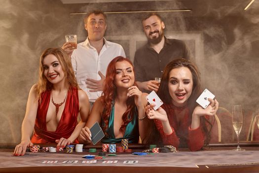Joyful buddies are playing poker at casino. They are celebrating their win, smiling and posing at the table against a dark smoke background. Cards, chips, money, alcohol, fortune, gambling, entertainment concept.