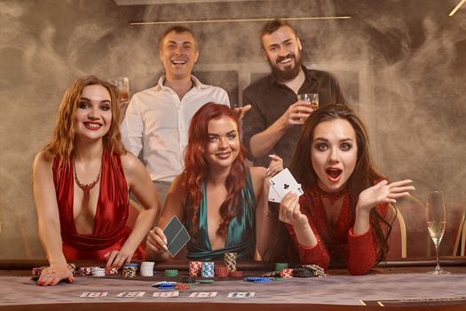 Joyful friends are playing poker at casino. They are celebrating their win, smiling and posing at the table against a dark smoke background. Cards, chips, money, alcohol, fortune, gambling, entertainment concept.