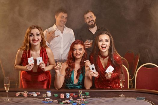 Group of a pleasant buddies are playing poker at casino. They are celebrating their win, looking at the camera and holding some playing cards in their hands while posing at the table against a smoke background. Chips, money, alcohol, gambling, entertainment concept.