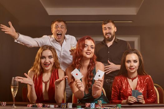 Happy buddies are playing poker at casino. They are celebrating their win, smiling and posing at the table against a dark smoke background. Cards, chips, money, alcohol, fortune, gambling, entertainment concept.