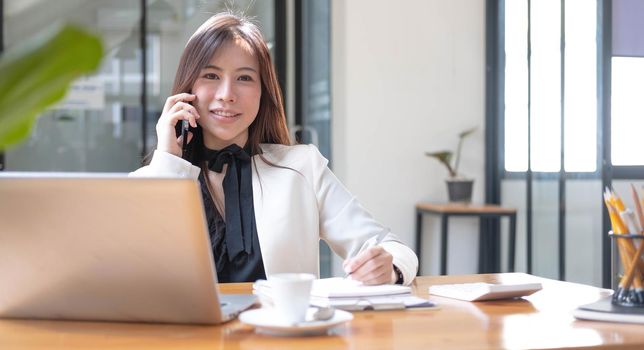 Charming Asian businesswoman working with a laptop and use smartphone at the office. Looking at camera..