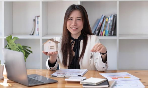 Miniature house in the hands of an Asian woman real estate agent home loan working at the office. Looking at the camera..