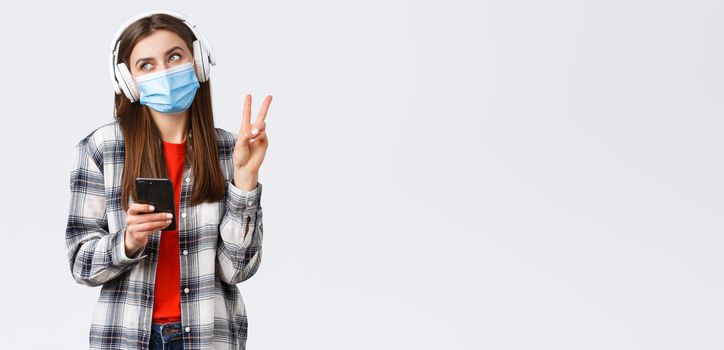 Social distancing, leisure and lifestyle on covid-19 outbreak, coronavirus concept. Happy cheerful silly girl in medical mask and headphones, show peace sign smiling, holding mobile phone.